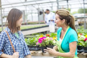 A garden center employee in a green apron selling flowers to an Asian woman in a flannel shirt.