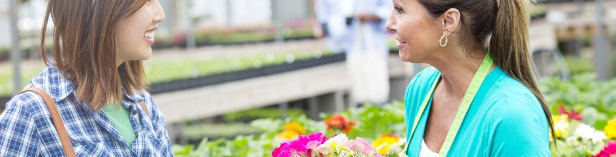 A garden center employee in a green apron selling flowers to an Asian woman in a flannel shirt.