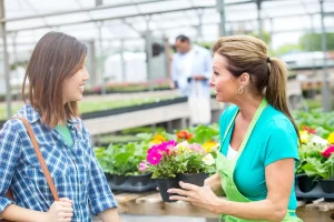 A garden center employee in a green apron selling flowers to an Asian woman in a flannel shirt.