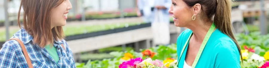 A garden center employee in a green apron selling flowers to an Asian woman in a flannel shirt.