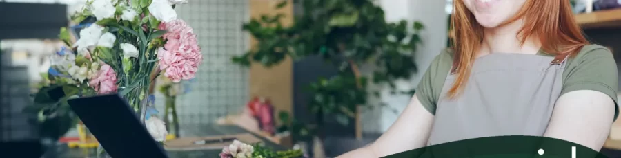 A smiling garden center employee stands behind a counter with flowers, using a touch screen monitor. It is set against a blurred background of a modern, green-themed space. Text: "Advancing Your Touchscreen Configuration" -- A Rapid Garden POS webinar