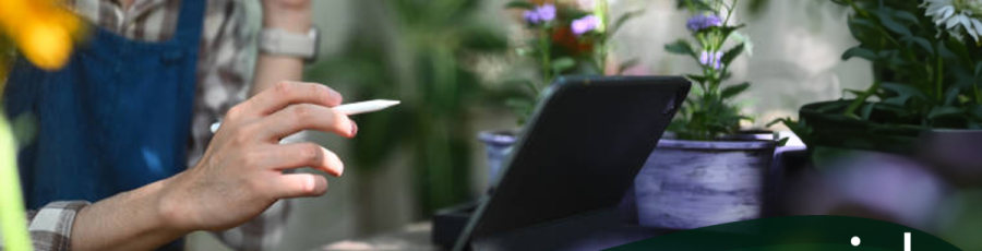 A man uses a tablet to create a sales kit within the robust Rapid Garden POS software in a garden retail setting. He is holding a pen and speaking on a phone while looking at a tablet. Surrounding them are colorful potted plants. Text: "Creating and Working with Sales Kits" -- a Rapid Garden POS webinar