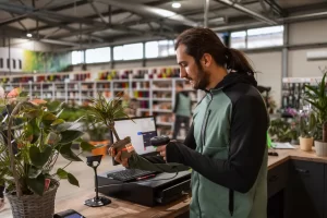 A man behind the cashier stand is scanning a price tag on a potted plant with a bar code reader.