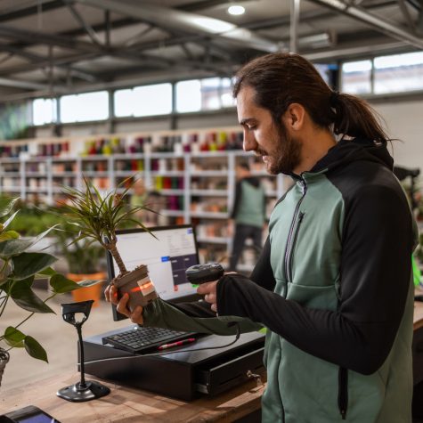 A man behind the cashier stand is scanning a price tag on a potted plant with a bar code reader.