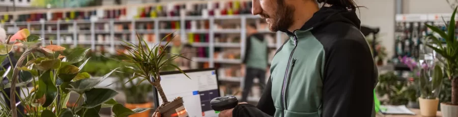 A man behind the cashier stand is scanning a price tag on a potted plant with a bar code reader.