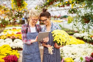 Two women in a garden center; one is using a tablet while the other is holding a potted plant.