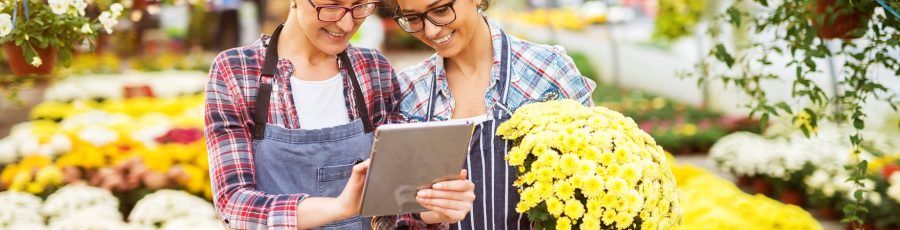 Two women in a garden center; one is using a tablet while the other is holding a potted plant.