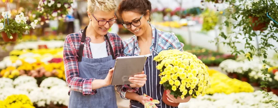 Two women in a garden center; one is using a tablet while the other is holding a potted plant.