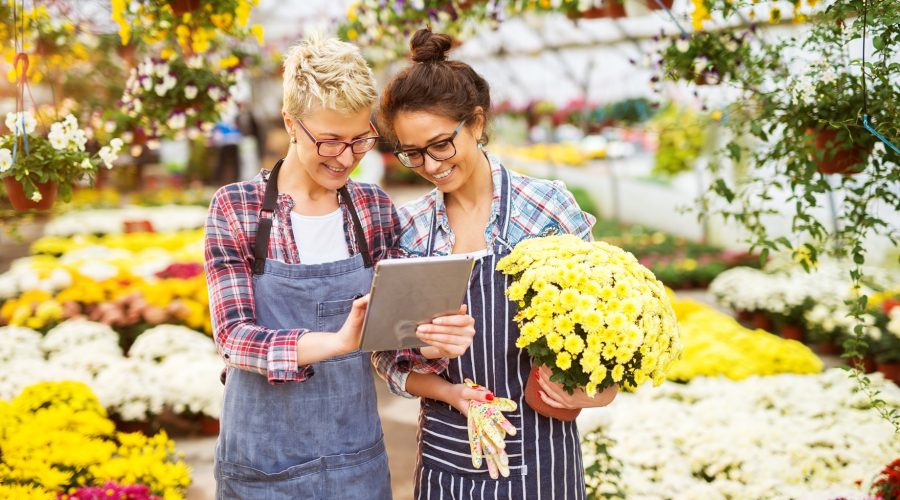 Two women in a garden center; one is using a tablet while the other is holding a potted plant.